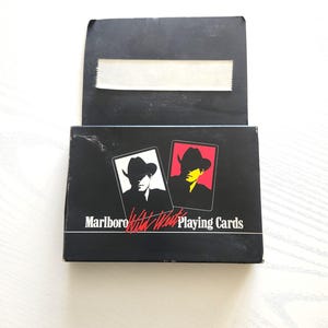 May include: Black playing card box with the text "Marlboro" and "What West Playing Cards". The box features two playing card designs with a silhouette of a person wearing a cowboy hat. One card is black and white, and the other is red and yellow.