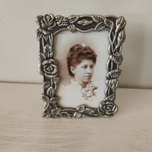 May include: A silver-colored, rectangular picture frame with a floral design. The frame surrounds a black and white photograph of a woman. The frame is decorated with roses and leaves.