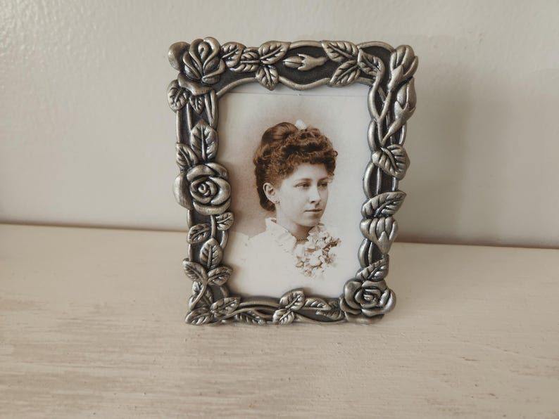 May include: A silver-colored, rectangular picture frame with a floral design. The frame surrounds a black and white photograph of a woman. The frame is decorated with roses and leaves.