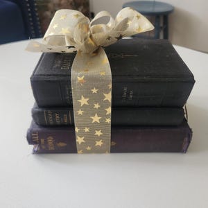 May include: A stack of three hardcover books tied together with a translucent beige ribbon featuring a gold star pattern. The top book is black with the text "DAILY LIGHT" visible on the spine. The bottom book is dark purple with the text "CALL OF THE BLOOD" visible on the spine.
