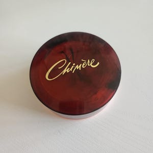 May include: A red and black round container with a gold inscription that reads "Chimère".