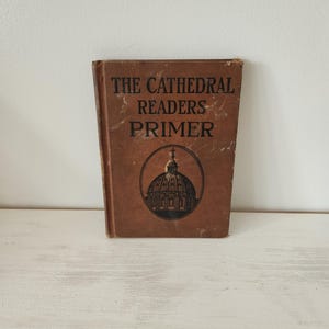 May include: A vintage book titled "The Cathedral Readers Primer" with a brown cover. The book features black text and a circular illustration of a cathedral. The book is standing upright on a white surface.