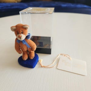May include: A small, brown teddy bear with a blue bow tie stands on a blue heart-shaped base. The bear is displayed next to a clear acrylic box with a black base. A tag is attached with a gold string.