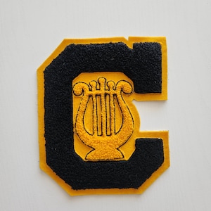 May include: A varsity letter patch in the shape of the letter "C". The patch has a black, textured background with a gold border. Inside the "C" is a gold lyre symbol.