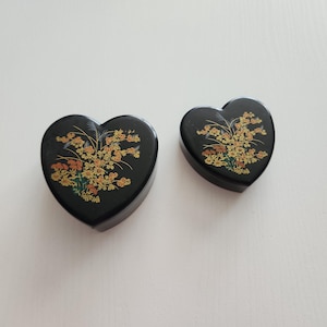 May include: Two heart-shaped black boxes with floral designs in yellow, orange, and green. The boxes are of different sizes, with the larger one in the foreground and the smaller one behind it. The floral design is centered on the top of each box.