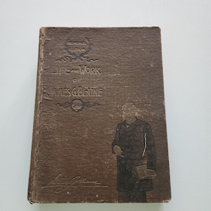 May include: A brown, antique book titled "Life and Work of James G. Blaine." The cover features embossed text and an illustration of a man. The book is a memorial edition.
