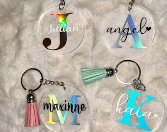 May include: Four clear acrylic keyrings with different coloured iridescent backgrounds. Each keyring has a letter charm with a name written below it. The names are Jillian, Angel, Maxine, and Kaia. Each keyring has a different coloured tassel.
