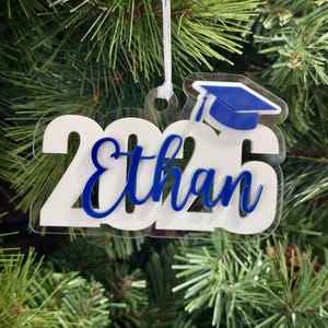 Graduation Christmas Ornament personalized with Students name in the color of the customers choice. 3 layer ornament has a clear background, white year and then the colored details like name and mortar board. Ornament hangs on a white ribbon.