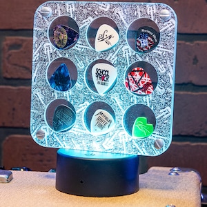 May include: A square, illuminated display case for guitar picks. The case is silver with a patterned background and holds nine guitar picks of various colors and designs. The base is black and sits on a textured surface.