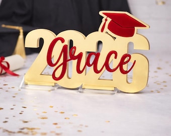 Puede incluir: Decoración de graduación dorada con la palabra "Grace" en escritura roja y el año "2020". Un birrete rojo se encuentra encima del "0". La decoración está sobre un soporte transparente, con una toga de graduación en el fondo.