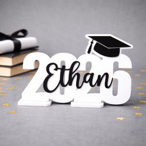 May include: A white wooden graduation decoration with the name "Ethan" in black script and the year "2020". A black graduation cap sits atop the "2020". Gold star confetti is scattered around the base.