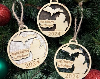 May include: Three wooden Christmas ornaments with the state of Michigan cut out. Each ornament has the text "First Christmas in Michigan 2024". The ornaments are hanging on a Christmas tree with red and green ornaments.