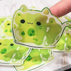 May include: A set of clear vinyl stickers featuring a cartoon green cat with a skull and crossbones design. The cat is depicted as a blob of goo with a cute face and a tail.