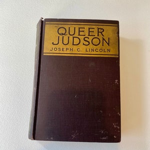 Könnte beinhalten: Ein Vintage-Hardcover-Buch mit dem Titel "Queer Judson" von Joseph C. Lincoln. Das Buch hat einen dunkelbraunen Einband mit einem goldenen rechteckigen Etikett oben. Titel und Autorenname sind in Schwarz gedruckt.