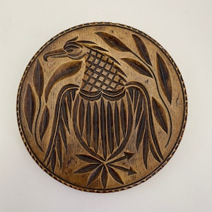 Könnte beinhalten: Eine runde Holzschnitzerei mit einem detaillierten Vogeldesign. Der Vogel hat einen langen Schnabel und ist von Blättern umgeben. Die Schnitzerei hat eine warme braune Farbe und eine strukturierte Oberfläche. Das Design ist in das Holz geschnitzt und erzeugt einen Relief-Effekt.