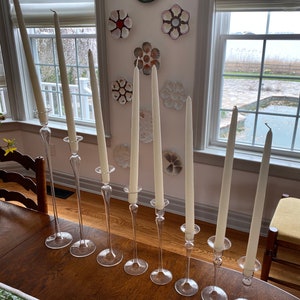 May include: Seven clear glass candle holders with long, white candles stand on a dark wooden table. The candle holders are displayed in a row. Decorative wall plates are visible in the background.