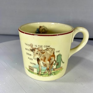 May include: A vintage cream ceramic mug with a red rim and a handle. The mug features a colourful illustration of a cow with text that reads "THIS IS THE COW WITH CRUMPLED HORN". A small figurine is inside the mug.