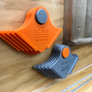 May include: Two fan-shaped artisan tools with magnetic fixings. One is orange and marked "1/2 Inch", the other is grey and marked "1/4 Inch". Both have "ARTISAN MADE" below the inch measurement. They are mounted on a wooden surface.
