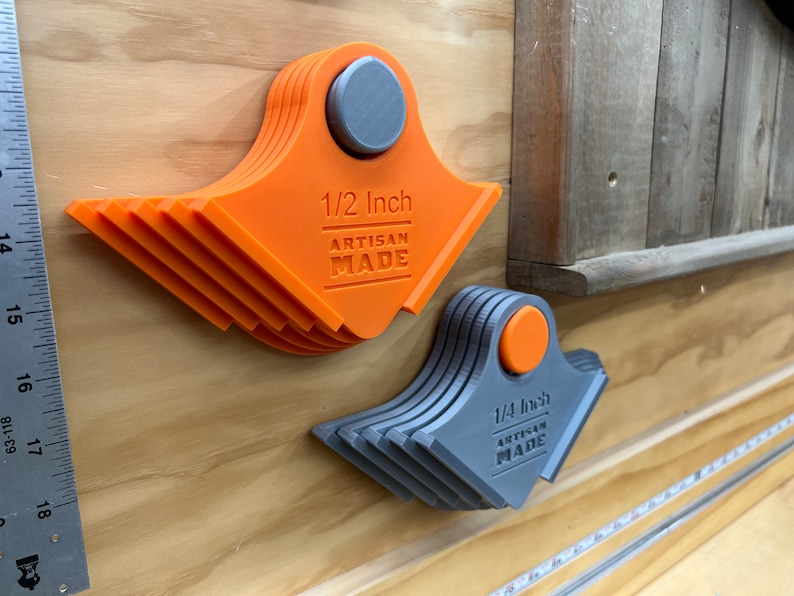 May include: Two fan-shaped artisan tools with magnetic fixings. One is orange and marked "1/2 Inch", the other is grey and marked "1/4 Inch". Both have "ARTISAN MADE" below the inch measurement. They are mounted on a wooden surface.