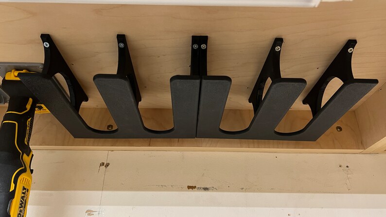 May include: Four black wall-mounted storage brackets with two slots each, designed to hold tools or other items. The brackets are mounted on a light wood surface.