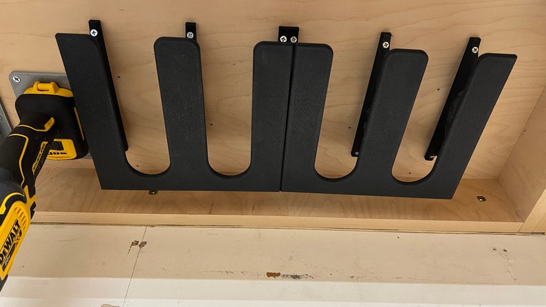 May include: Close-up of a set of four black plastic snowboard storage racks mounted inside a wooden cabinet. The racks are designed to hold snowboards vertically. A yellow and black cordless drill is visible in the foreground.