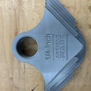 May include: A stack of grey 3D-printed tools with a circular opening and a notched edge. The text "1/4 Inch ARTISAN MADE" is printed on the tool. An orange tool is visible in the background. The tools are on a wooden surface.