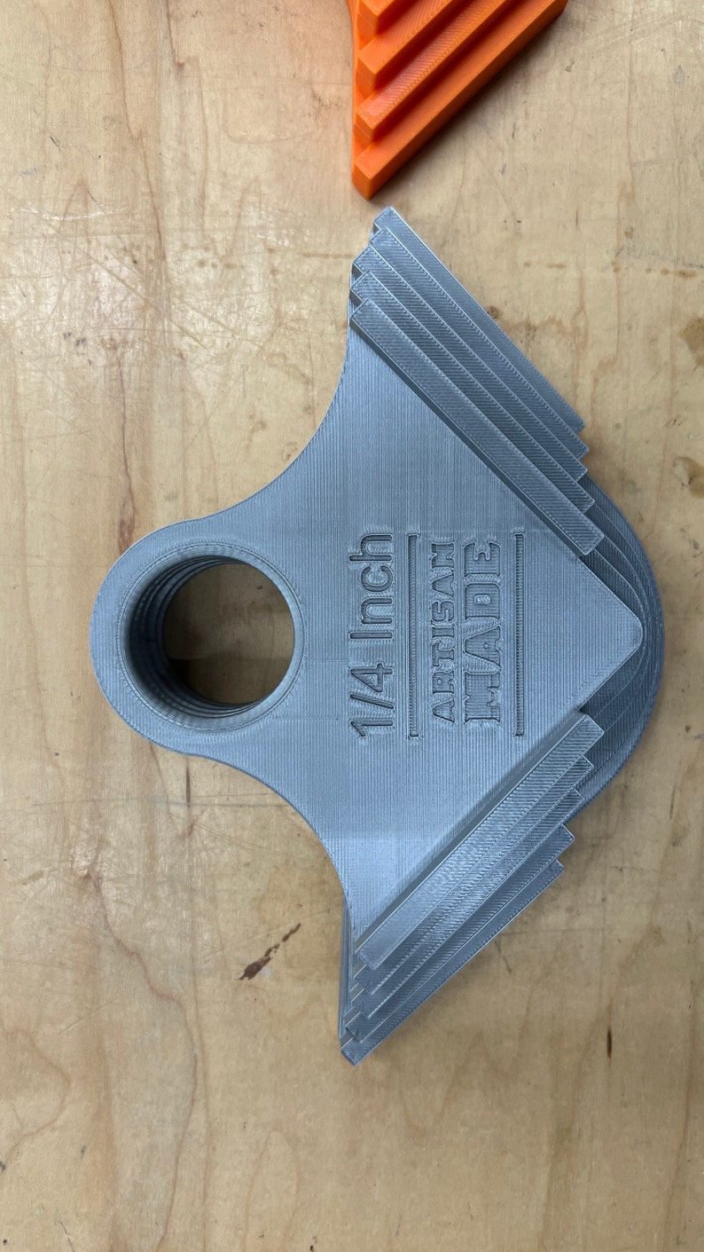 May include: A stack of grey 3D-printed tools with a circular opening and a notched edge. The text "1/4 Inch ARTISAN MADE" is printed on the tool. An orange tool is visible in the background. The tools are on a wooden surface.