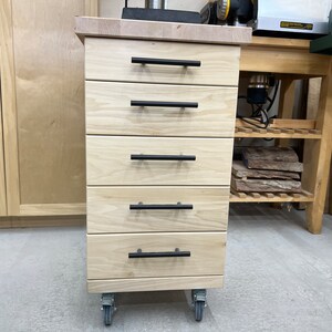May include: A light wood five-drawer rolling cabinet with black drawer pulls. The cabinet is on wheels and has a wood top.