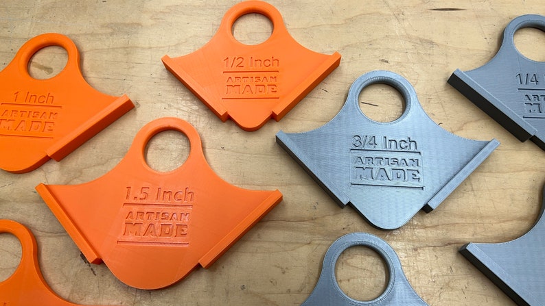 May include: A selection of orange and grey Artisan Made corner radius jigs. The jigs are labelled with measurements: 2.5 cm, 1.3 cm, 3.8 cm, 1.9 cm, and 0.6 cm. Each jig has a handle and a beveled edge.