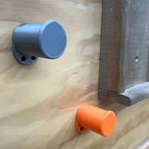 May include: Two cylindrical, grey and orange wall-mounted knobs are fixed to a light-coloured wooden surface. The knobs have a circular base with two holes for mounting. A section of weathered wood is visible in the background.