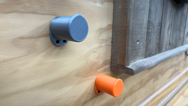 May include: Two cylindrical, grey and orange wall-mounted knobs are fixed to a light-coloured wooden surface. The knobs have a circular base with two holes for mounting. A section of weathered wood is visible in the background.