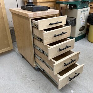 May include: A wooden cabinet with five drawers on wheels. The drawers are open and have black handles. The cabinet is made of light wood and has a light wood top.