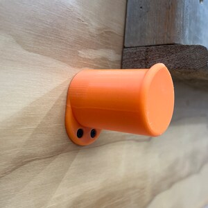 May include: An orange, cylindrical holder is mounted on a light-coloured wooden surface. The holder has a circular base with two black screw holes and a flared opening. The background includes a wooden ledge. The object is likely a wall-mounted accessory.