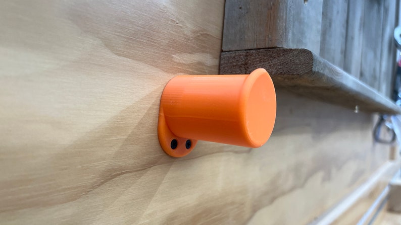 May include: An orange, cylindrical holder is mounted on a light-coloured wooden surface. The holder has a circular base with two black screw holes and a flared opening. The background includes a wooden ledge. The object is likely a wall-mounted accessory.