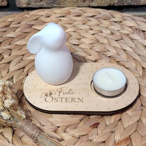 Könnte beinhalten: Eine weiße Keramik-Hasenfigur steht auf einem Holzsockel mit einer kleinen weißen Kerze in einem Glashalter. Der Sockel ist mit dem Text "Frohe Ostern" graviert.
