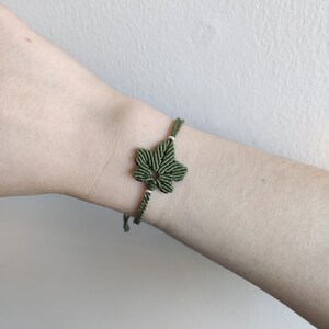 Può includere: Un braccialetto in macramè fatto a mano con un motivo a foglia verde. Il braccialetto è realizzato con cordoncino verde e bianco, con la foglia come punto focale. Il braccialetto è indossato su un polso su uno sfondo neutro.