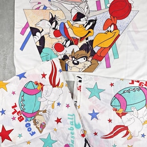 May include: White bedsheets featuring Looney Tunes characters. The top sheet has a triangular design with Bugs Bunny, Daffy Duck, Sylvester, and other characters. The pillowcases have football and star designs with Taz and Foghorn Leghorn.