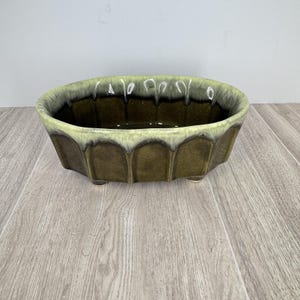May include: A vintage, oval-shaped planter with a ridged exterior. The planter is a deep olive green with a light green and white glazed rim. It has three small feet and is likely made of ceramic.