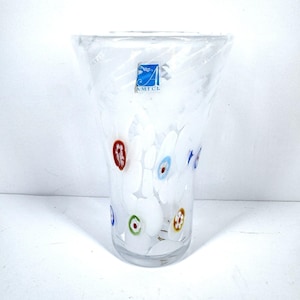 May include: A clear glass vase with white floral designs and colorful accents. The vase has a flared top and a cylindrical body. The floral designs include red, blue, green, and yellow details. The vase has a blue and white logo that says "AMICL".