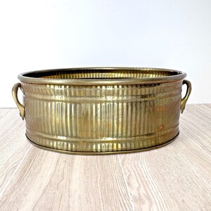 May include: A vintage brass oval tub with vertical ribbed sides and two curved handles. The tub has a warm, golden hue and shows signs of age, adding to its antique charm. It is a decorative piece, suitable for various uses.