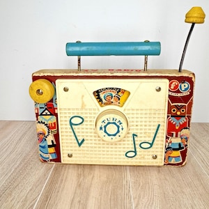 May include: Vintage Fisher-Price toy radio with a cream-colored front panel and a wooden handle. The radio features illustrations of Native American figures and musical notes. The dial reads "TURN". The radio has a yellow antenna and a red, white, and blue color scheme.