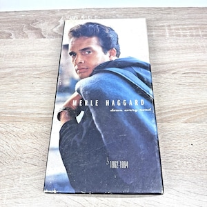 May include: A rectangular music box set featuring a portrait of Merle Haggard wearing a dark blue hooded jacket. The text reads MERLE HAGGARD down every road 1962-1994. The design is printed on a vertical cardboard sleeve with a smooth finish.