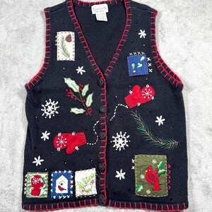 May include: A black knit vest featuring a V-neckline and red trim along the edges. The vest is decorated with embroidered patches and motifs including red mittens, snowflakes, holly leaves, a cardinal, and a pine branch. A label reads Basic Editions.