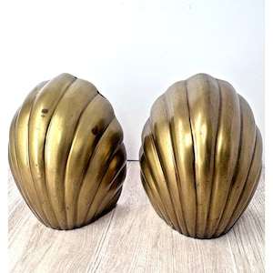 May include: Two golden-colored, shell-shaped decorative objects. Each shell has a ribbed texture and a rounded, curved form. The objects are placed side by side against a white background, suggesting a decorative or artistic theme.