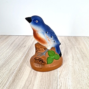 May include: A ceramic bluebird figurine perched on a brown tree stump base. The bird has a blue body, orange chest, and black beak. Green leaves and a small branch are also visible on the base. The figurine is a decorative item.