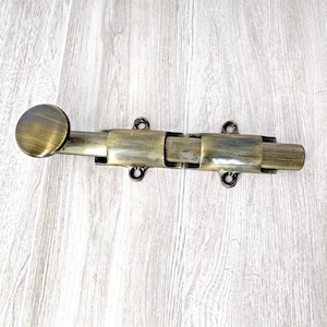 Puede incluir: Un cerrojo de barril de color bronce con un mango redondo y un cuerpo rectangular. El cerrojo tiene un mecanismo deslizante y está diseñado para asegurar puertas o portones. El herraje es de metal y tiene una estética vintage.