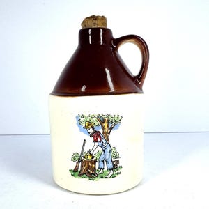 May include: A ceramic jug with a brown top and handle, and a cream-colored base. The base features a colorful illustration of a person with a rifle, a dog, and a tree. The jug is topped with a cork stopper.