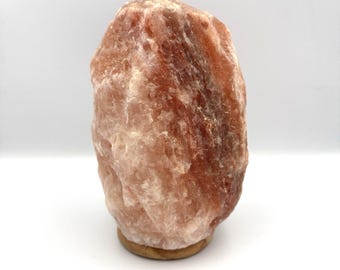 May include: A raw, crystalline Himalayan salt lamp with a rough, uneven texture and varied shades of translucent pink, orange, and deep red. The salt block is mounted on a circular, light-colored wooden base, creating a natural, organic appearance.