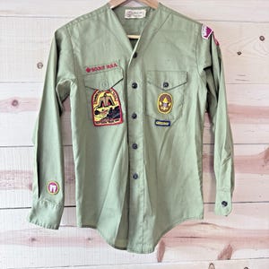 May include: A long-sleeve olive green button-down shirt featuring a V-neck collar, two chest pockets, and multiple embroidered patches. The patches include text reading SCOUT B.S.A., TANNEHILL IRON TRAIL 1965, and BIRMINGHAM, along with various scout emblems.