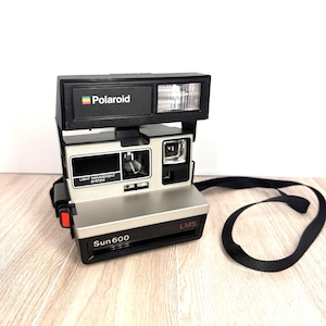 May include: A vintage Polaroid Sun 600 instant camera. The camera is black and silver with a built-in flash and a black strap. The Polaroid logo is visible on the top of the camera.